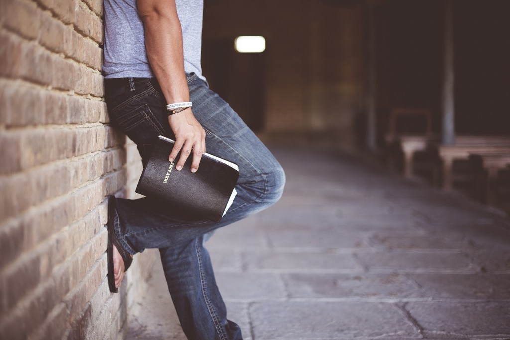 A man leaning against a wall holding a Bible, dressed in casual attire with jeans and a T-shirt.