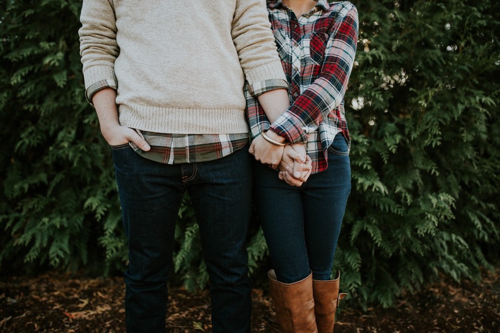 A young couple standing close together, holding hands, with a green bush in the background. The man is wearing a beige sweater over a plaid shirt, while the woman is dressed in a red and black plaid shirt and brown boots.