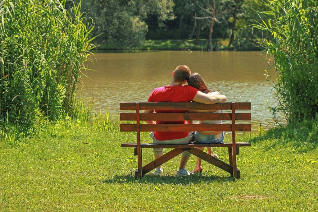 A couple sitting together on a wooden bench by a lake, enjoying each other's company amidst greenery.