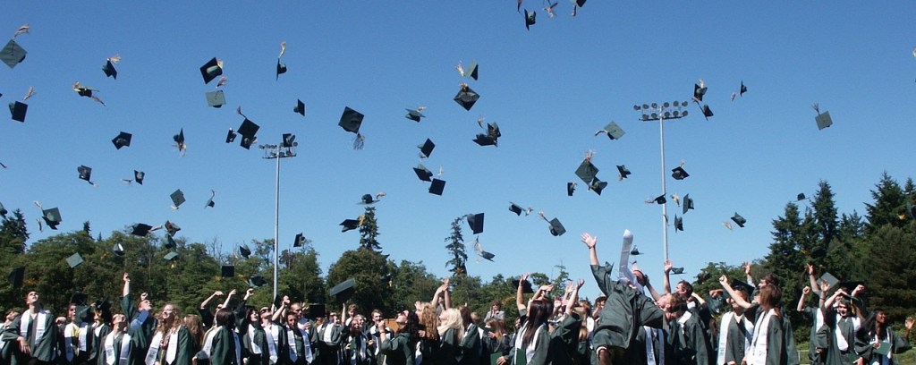 Graduating high school seniors joyfully throw their caps in the air during a graduation ceremony, celebrating their achievement against a clear blue sky.