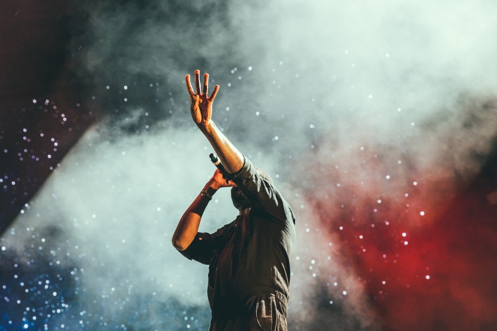 A performer singing passionately on stage with a raised hand, surrounded by a smoky atmosphere and colorful lights.