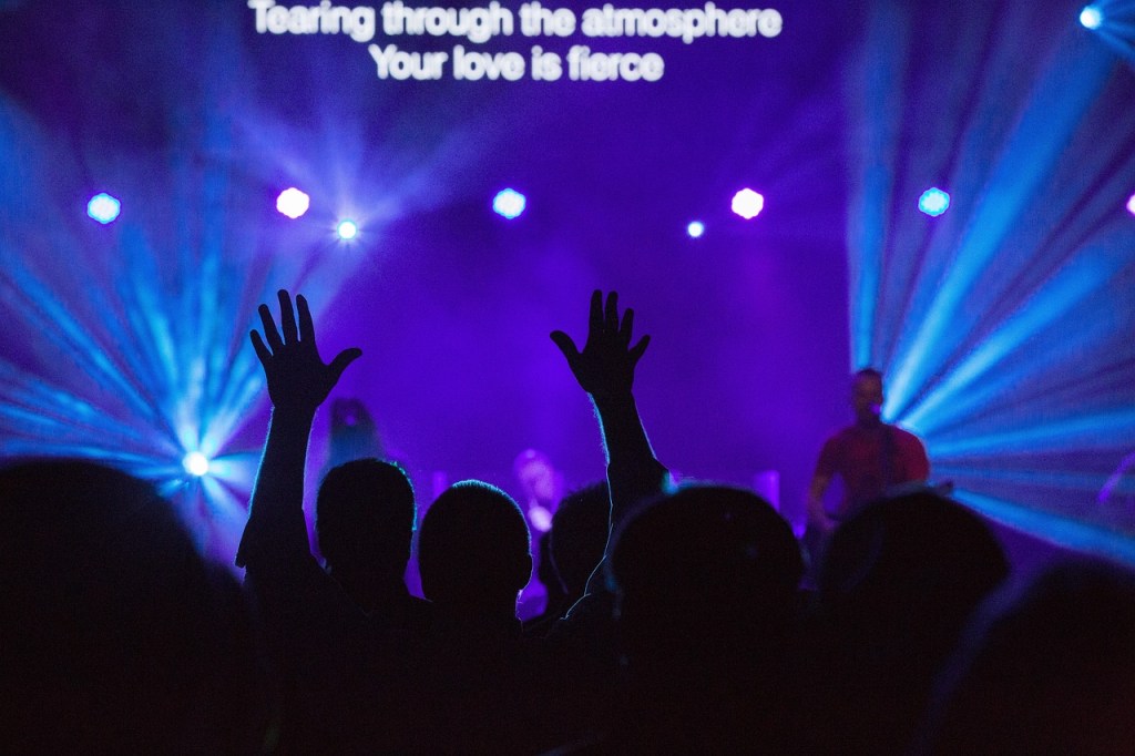 A crowd raising their hands in worship during a contemporary Christian music performance, with colorful stage lights and lyrics projected in the background.