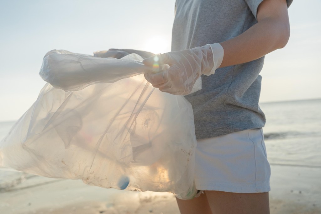 Person wearing gloves holding a transparent bag filled with trash on a beach, with the sun shining in the background.