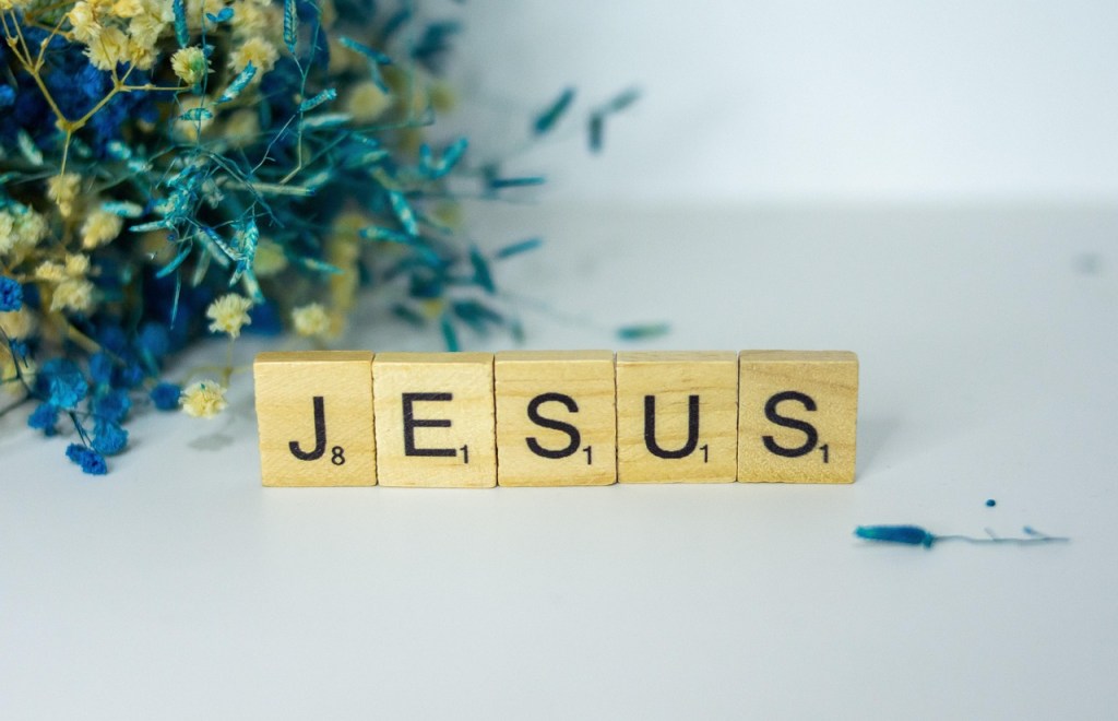 Wooden letter blocks spelling out 'JESUS' on a white surface with dried flowers in the background.