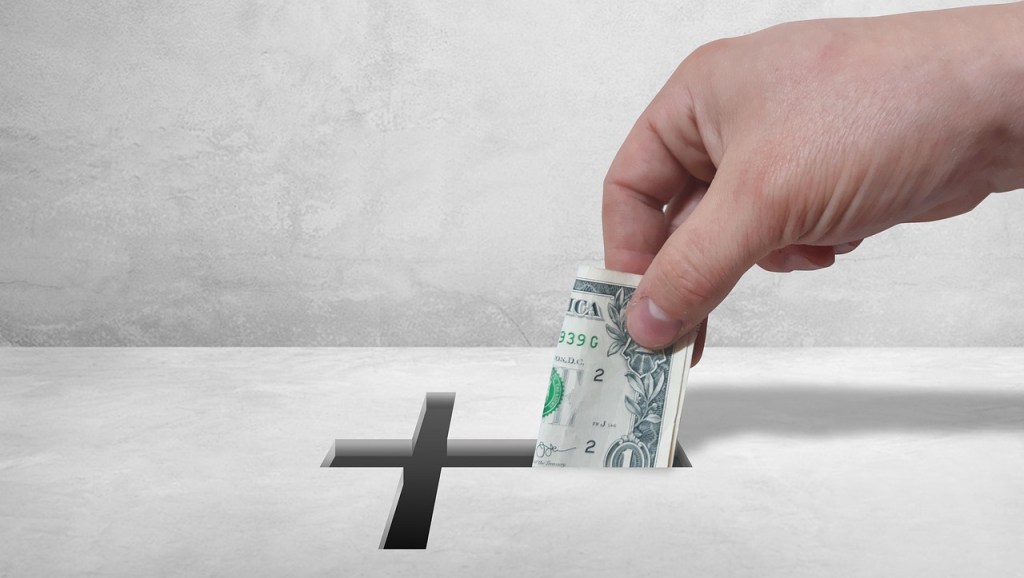 A hand inserting a dollar bill into a donation slot shaped like a cross on a gray background.