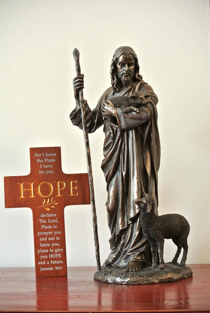 A bronze statue of Jesus as the Good Shepherd holding a lamb in His arms, accompanied by a wooden cross displaying the word 'HOPE' and a Bible verse.
