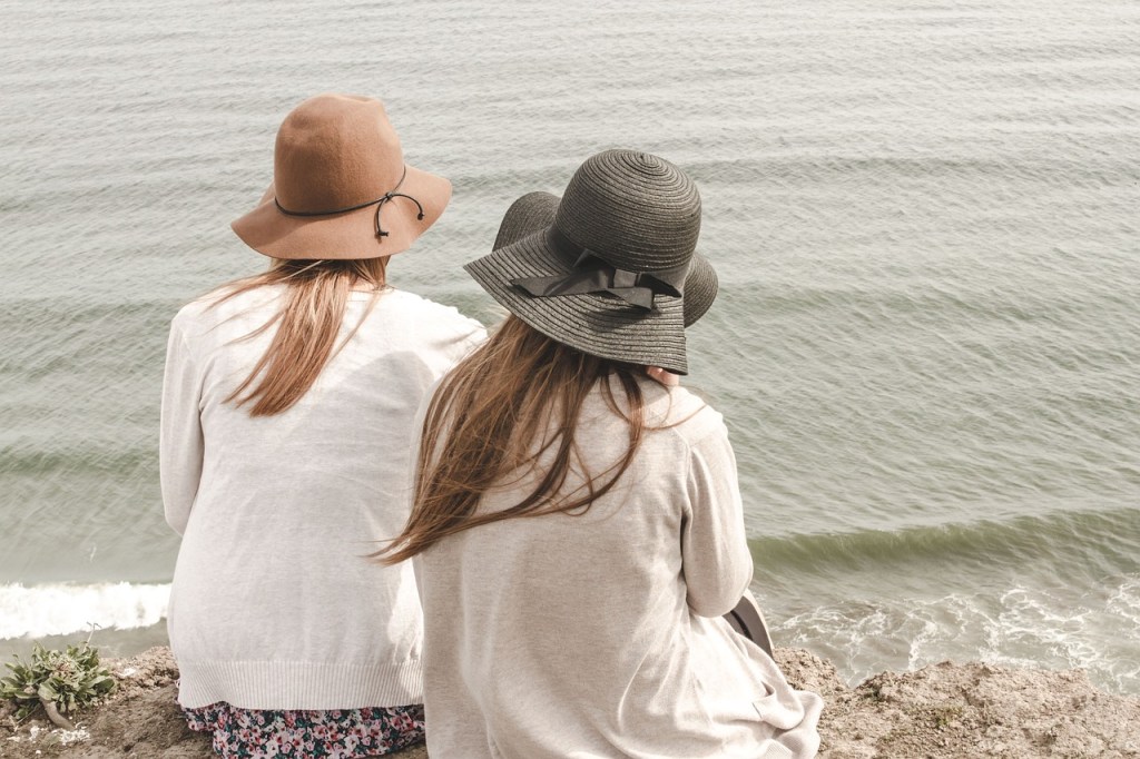 Two women sitting on a cliff overlooking the ocean, wearing hats and casual clothing.