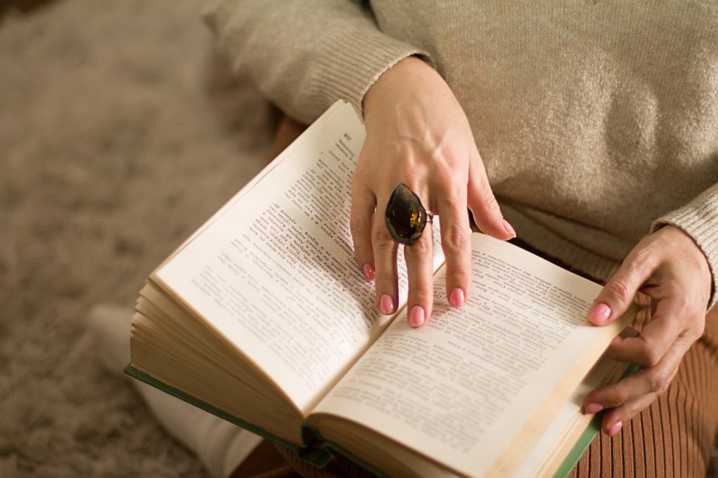 A person's hand holding open a book with a large, decorative ring on their finger, sitting on a soft, textured surface.