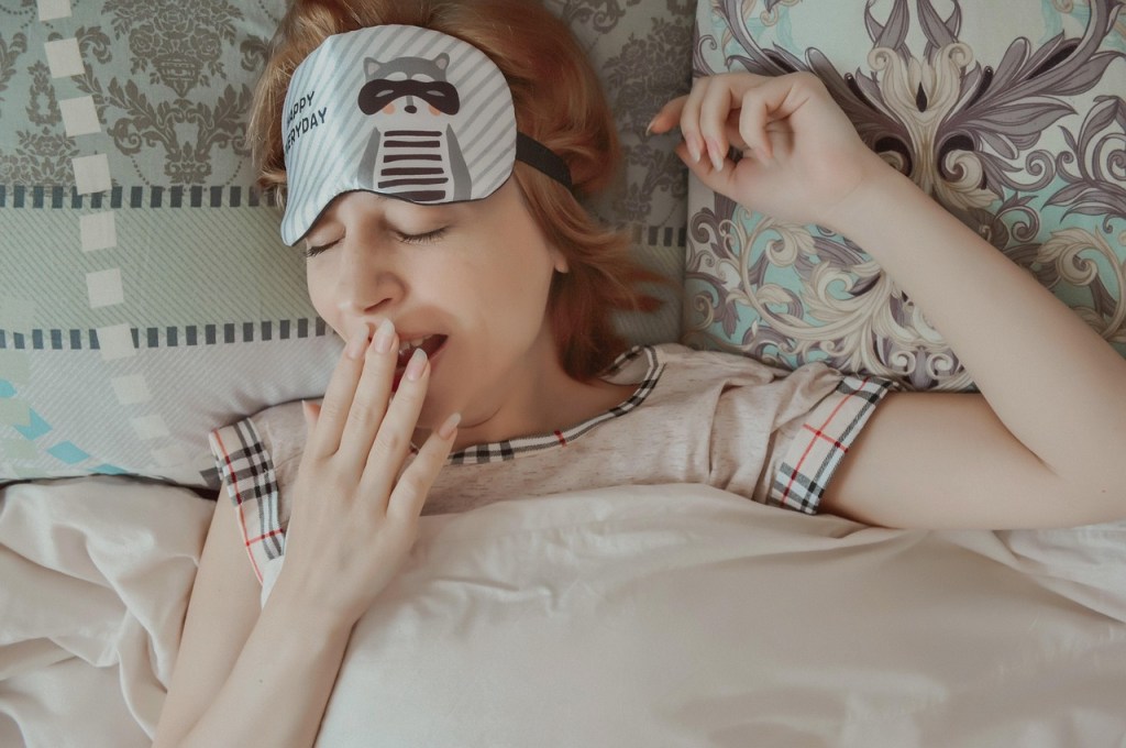 A person wearing a sleep mask yawns in bed, surrounded by colorful pillows.