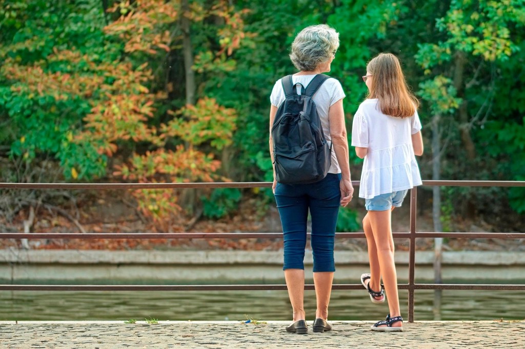 A grandmother and a young girl standing together by a railing with a green forest in the background, symbolizing mentorship and intergenerational connection.