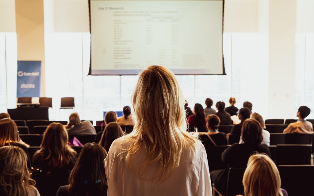 A woman with long blonde hair facing a projector screen in a conference room, where an audience of women is seated.