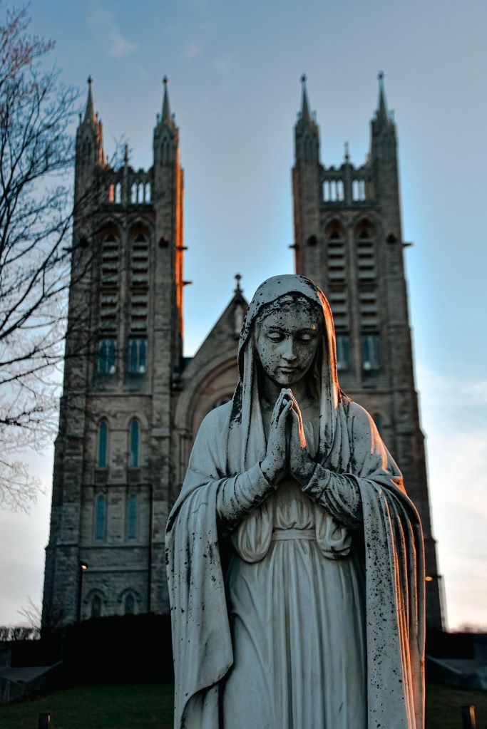 A statue of a woman with folded hands in prayer, positioned in front of a tall stone church with two prominent towers.