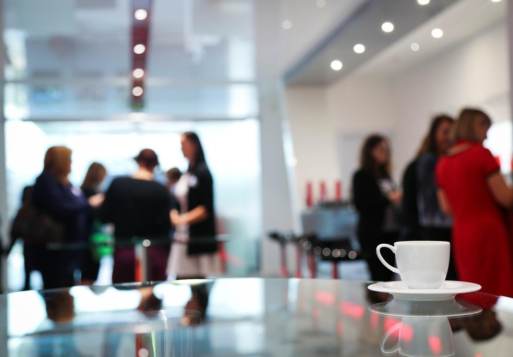 A blurred background of a group of women conversing at an event, with a focus on a white coffee cup and saucer placed on a glass table in the foreground.
