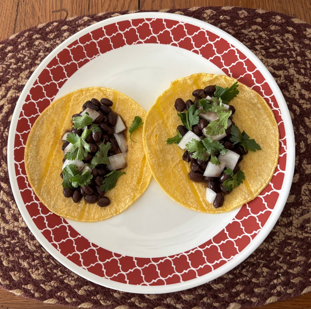 Two plant-based tacos on a decorative white and red plate, filled with black beans, diced onions, and fresh cilantro, resting on a woven placemat.