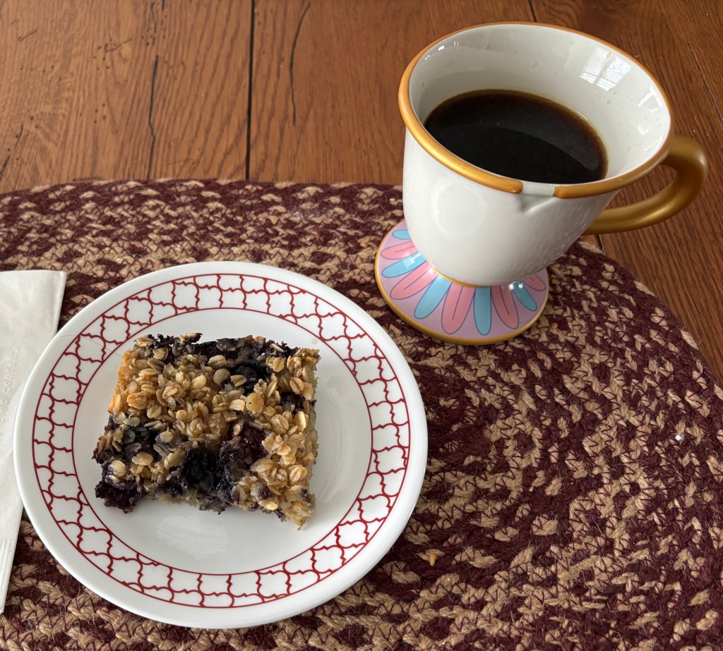 A plate of baked oatmeal with blueberries sits next to a cup of black coffee, all placed on a woven table mat.