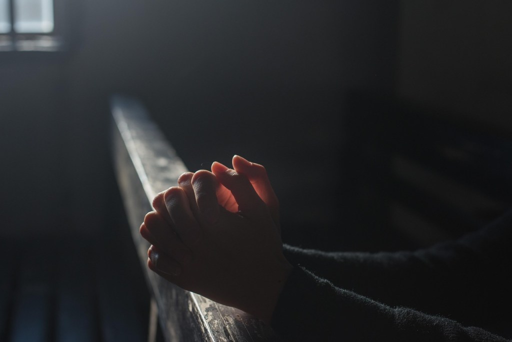 A person's hands resting on a wooden surface, symbolizing quiet prayer and reflection in a dimly lit space.