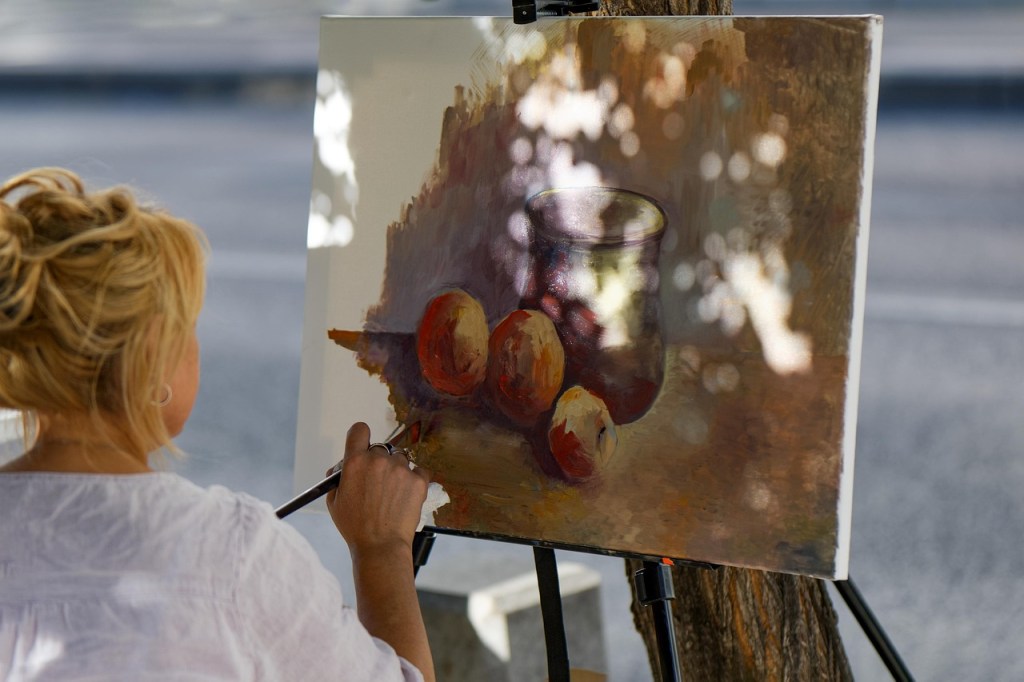 A woman with blonde hair paints a still life featuring three apples and a jar on a canvas set on an easel, with a blurred outdoor background.