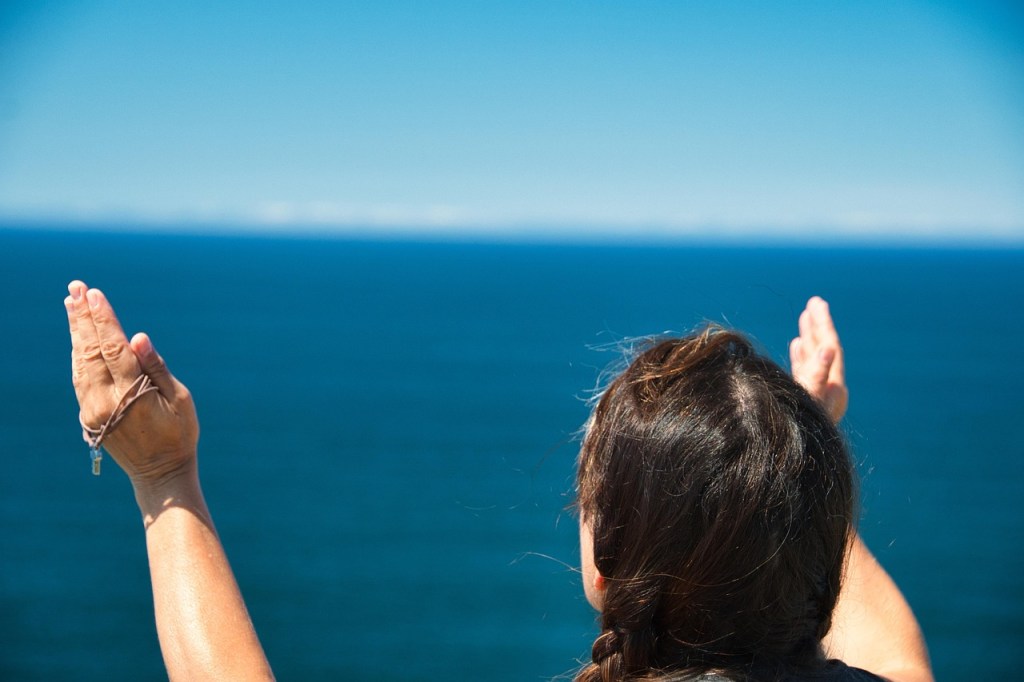 A woman with long hair, seen from behind, raises her hands in front of a vast ocean under a clear blue sky.