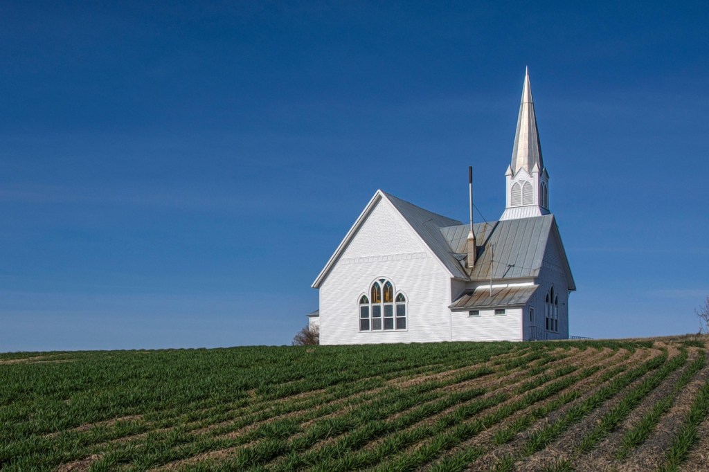 A white church with a tall steeple situated on a green field under a blue sky.
