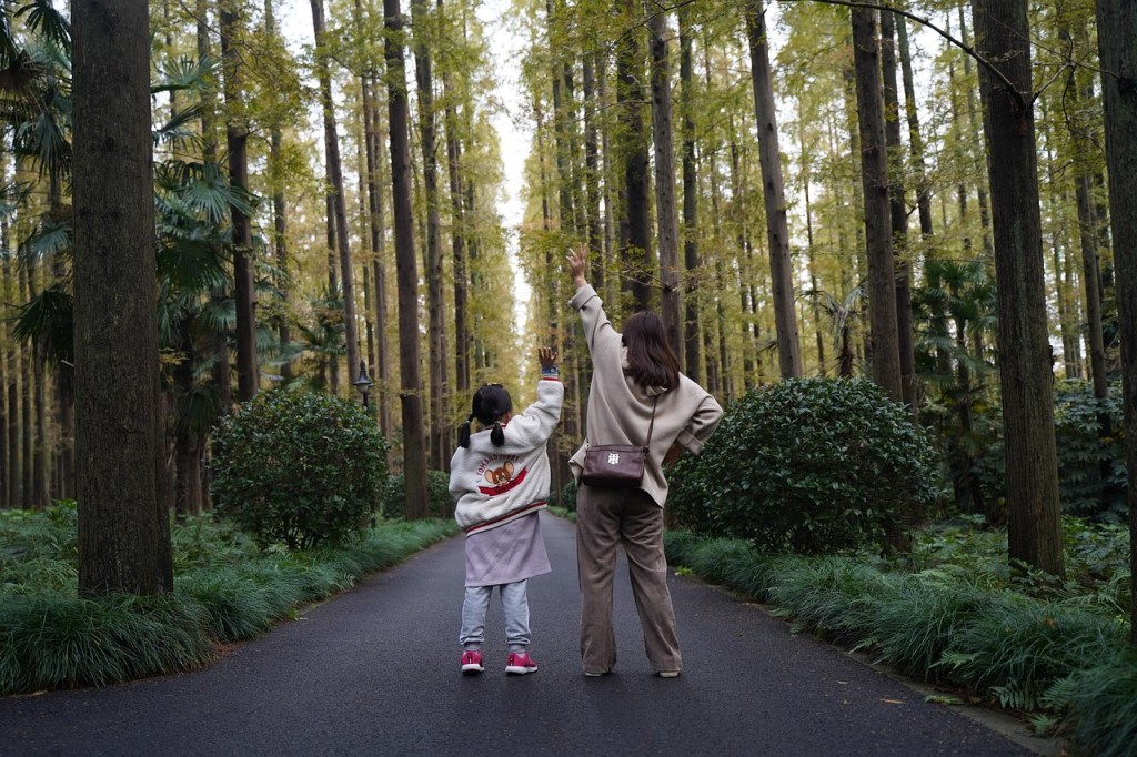 A woman and a girl standing on a path in a forest, both raising one hand in the air, surrounded by tall trees and greenery.