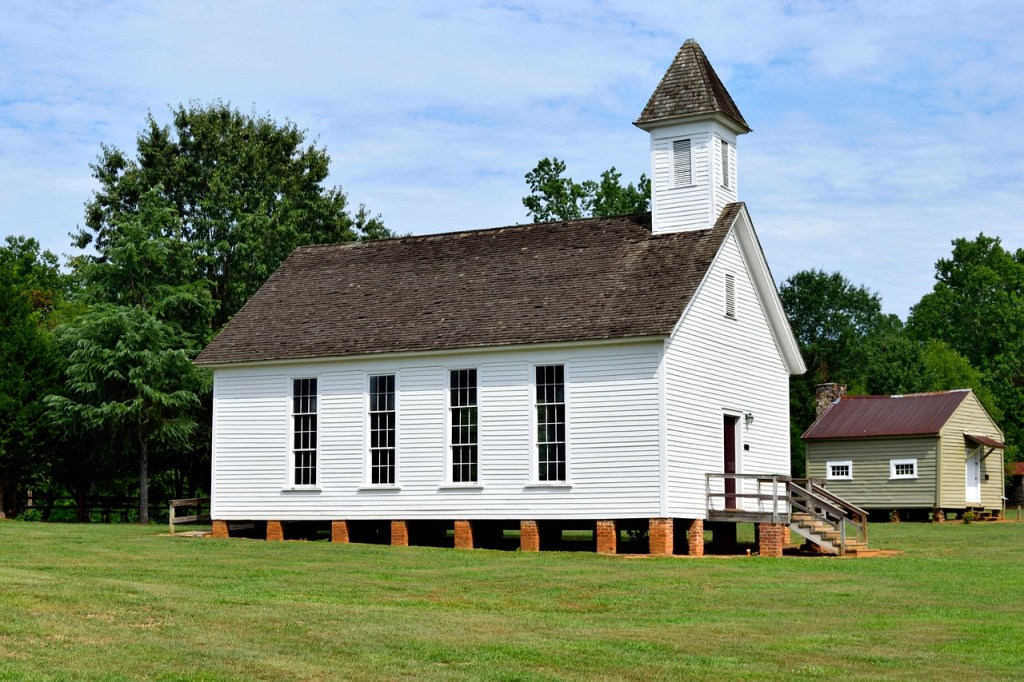 A white church building with a steep roof and a bell tower, surrounded by green grass and trees.