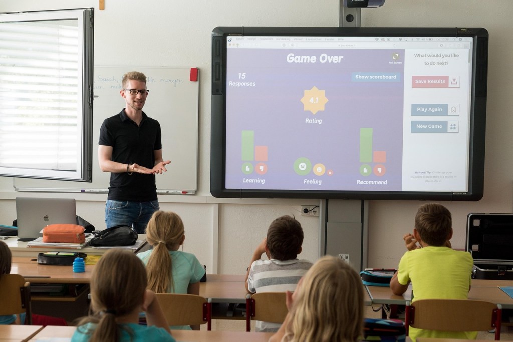 A teacher presenting and interacting with students in a classroom, with a digital scoreboard displaying game results on a projector.