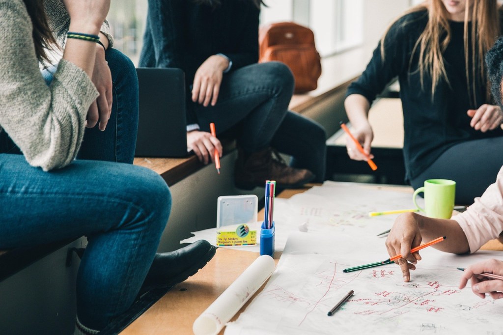 A group of students engaged in a collaborative study session, sitting around a table covered with papers, while holding pens and markers.