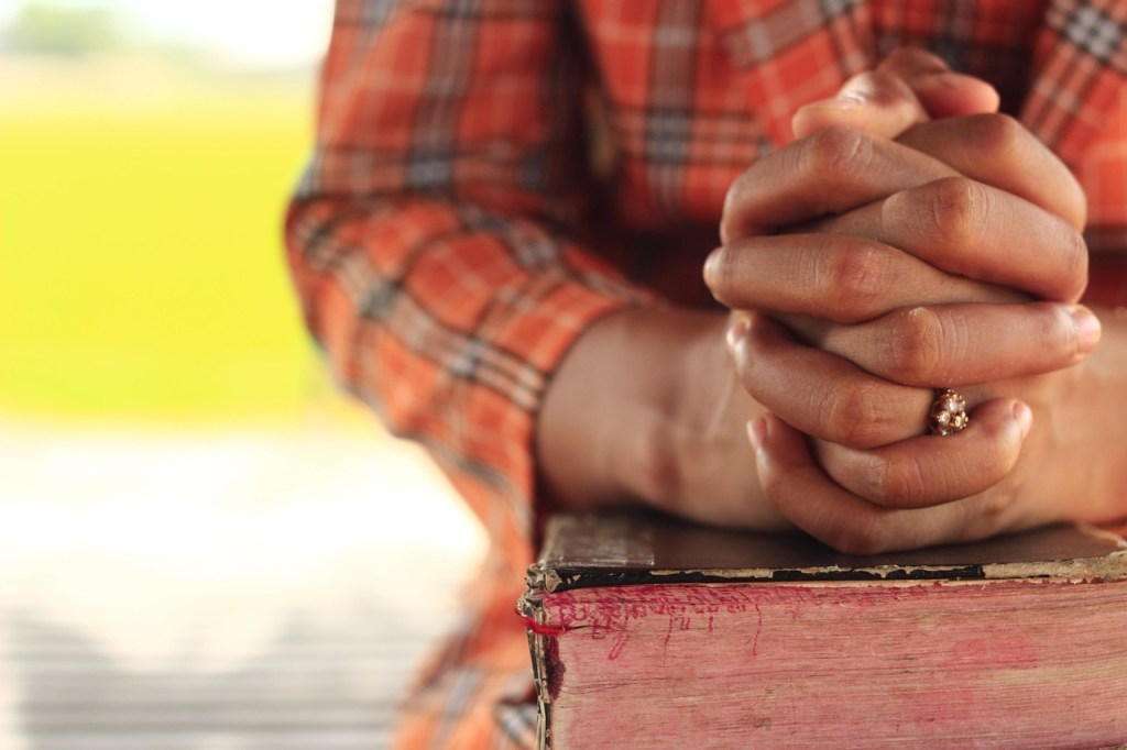 A person with clasped hands in prayer resting on a book, wearing an orange plaid shirt.