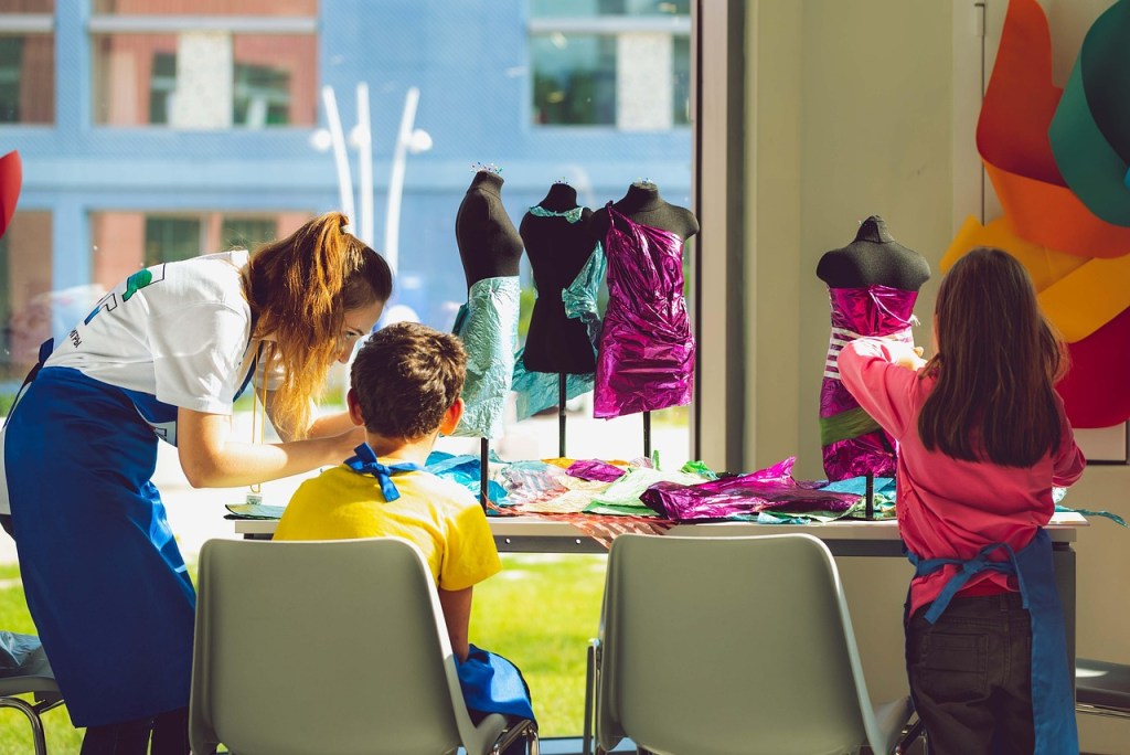 A teacher assists young students in an art project, creating colorful designs on dress forms in a bright classroom with large windows.