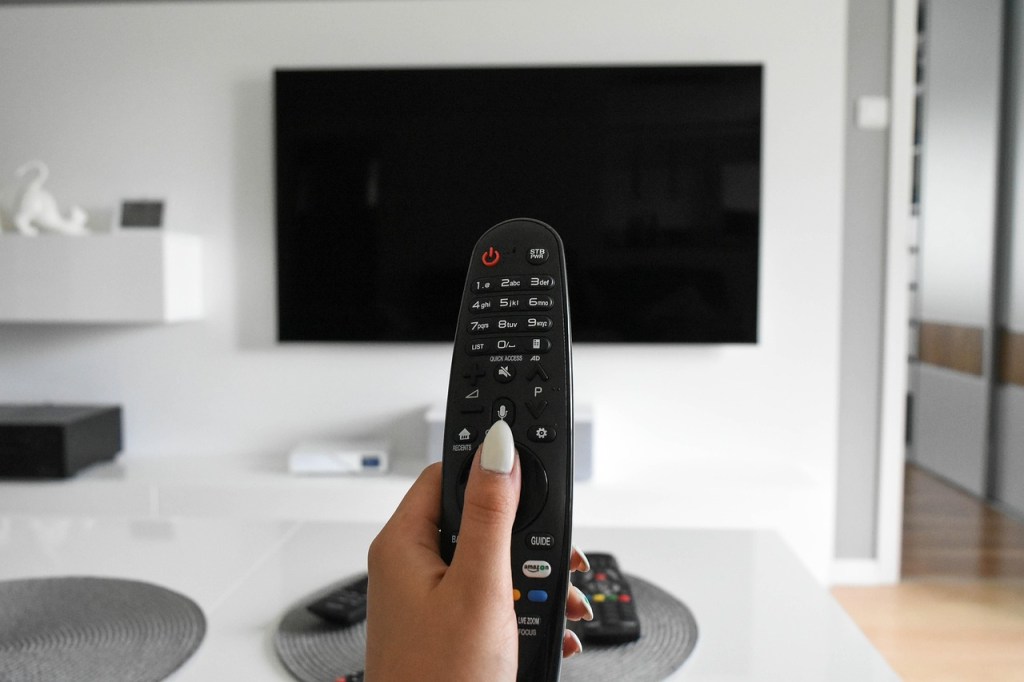 A person holding a TV remote control in front of a large, turned-off flat-screen television in a modern living room.