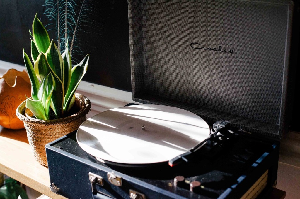 A close-up of a record player with a white vinyl record on it, alongside a potted green plant and a decorative pot in a warm, sunlit setting.