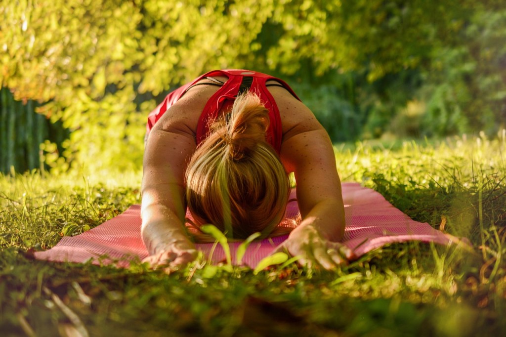 A person practicing yoga in a tranquil outdoor setting, stretching forward on a pink mat surrounded by greenery.