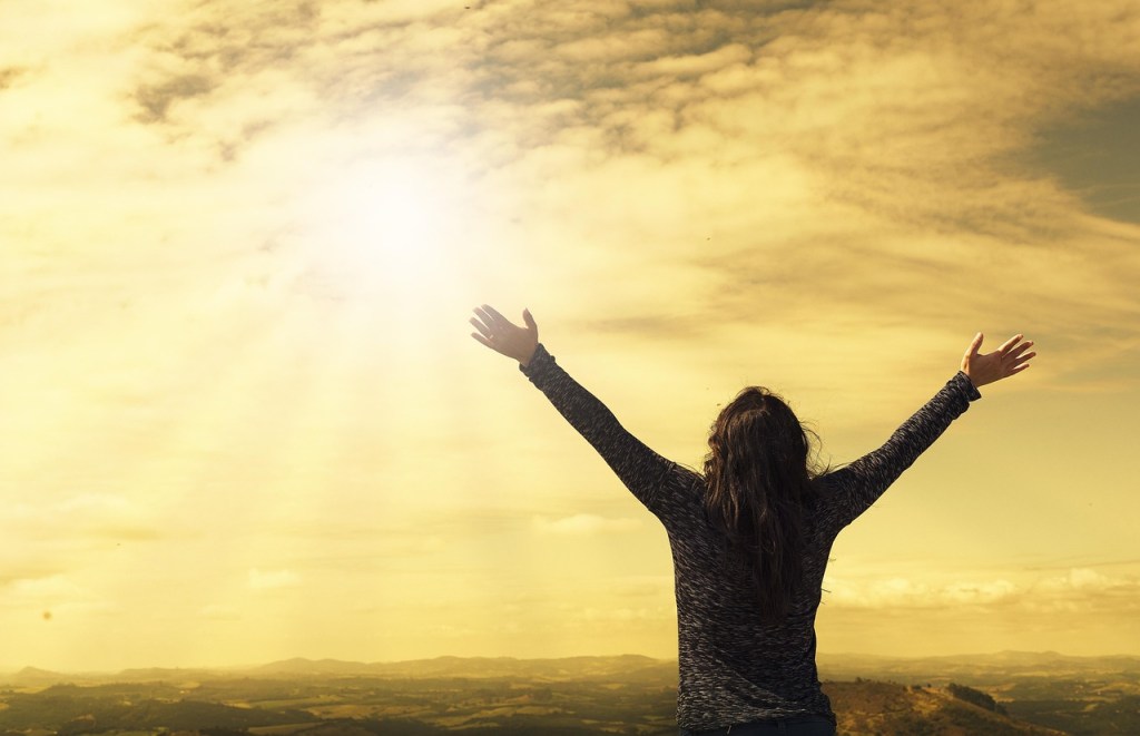 A person standing on a hill with arms raised towards a bright sun in a cloudy sky.