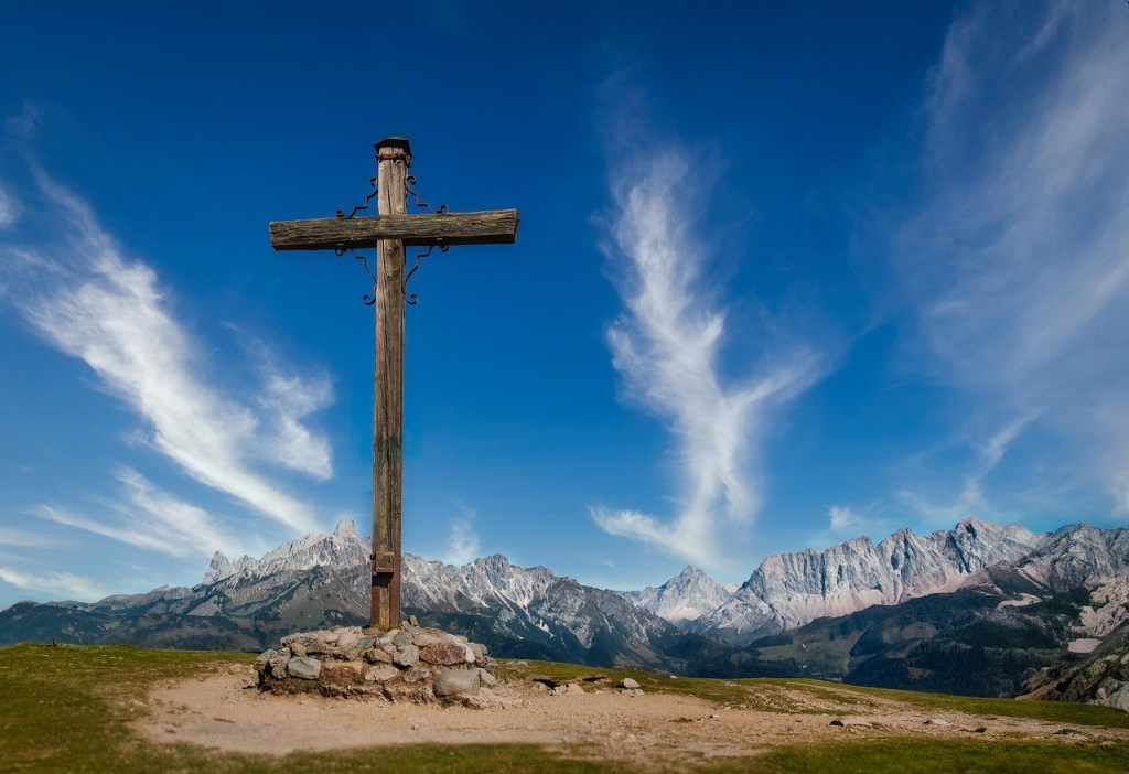 A wooden cross stands on a rocky base against a backdrop of mountains and a blue sky with wispy clouds.