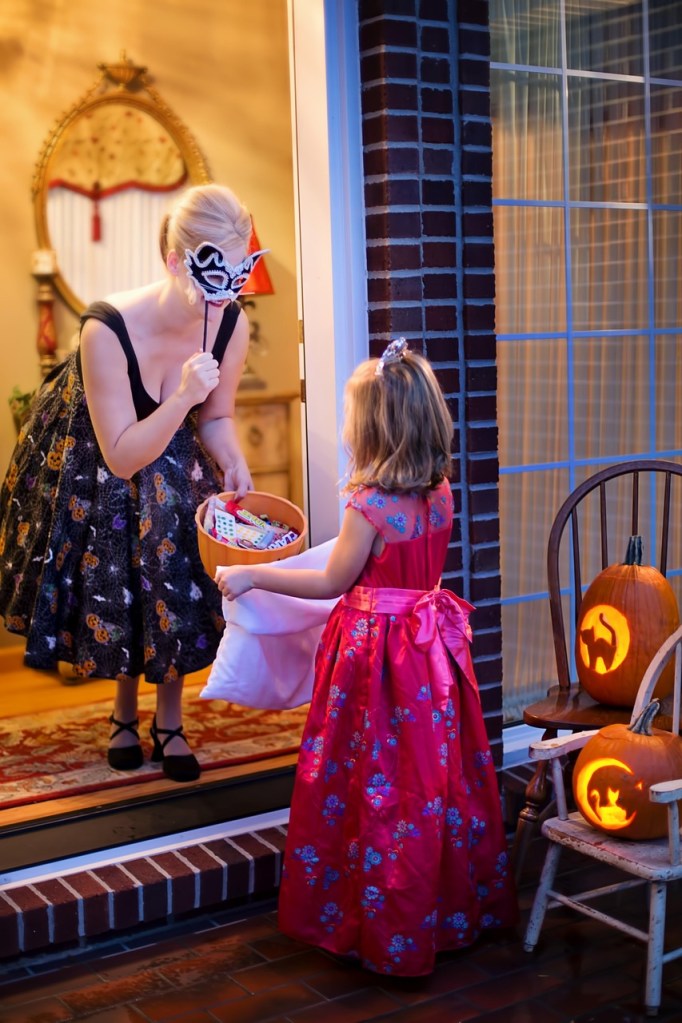 A woman wearing a masquerade mask greets a young girl in a red dress at a decorated porch during Halloween, with glowing jack-o'-lanterns in the background.
