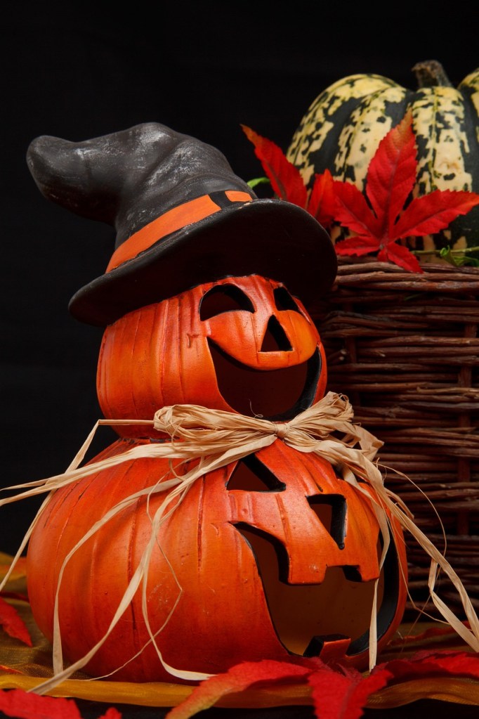 Two carved pumpkins stacked together, wearing a black witch hat, and adorned with a bow made of straw. A decorative basket and colorful autumn leaves are in the background.