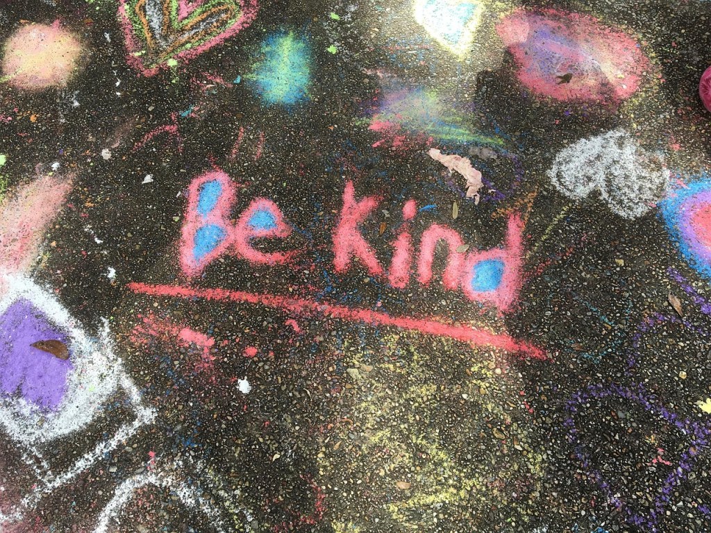 Colorful chalk art on pavement featuring the phrase 'Be Kind' in pink surrounded by other chalk drawings.