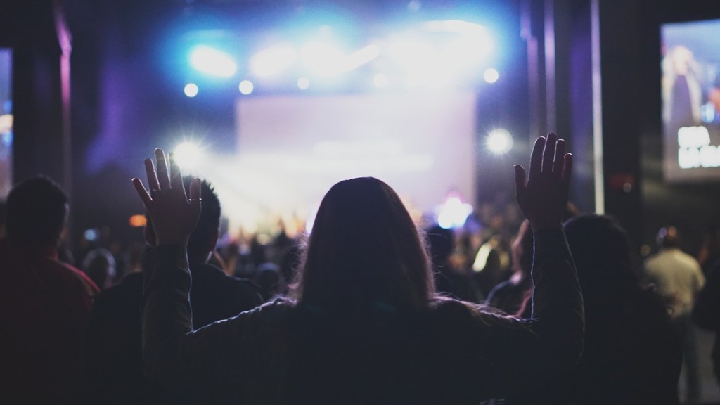 A crowd of worshippers with hands raised high in praise, illuminated by bright stage lights.