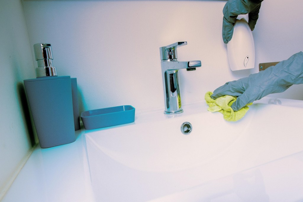 A person wearing gloves wipes down a sink with a cloth, showcasing a cleaning process. The sink area features a chrome faucet, a soap dispenser, and a small tray.