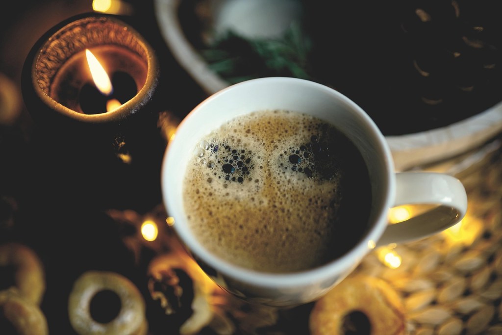 A close-up of a coffee cup filled with frothy coffee, next to a lit candle and surrounding decor, conveying a cozy and peaceful atmosphere.