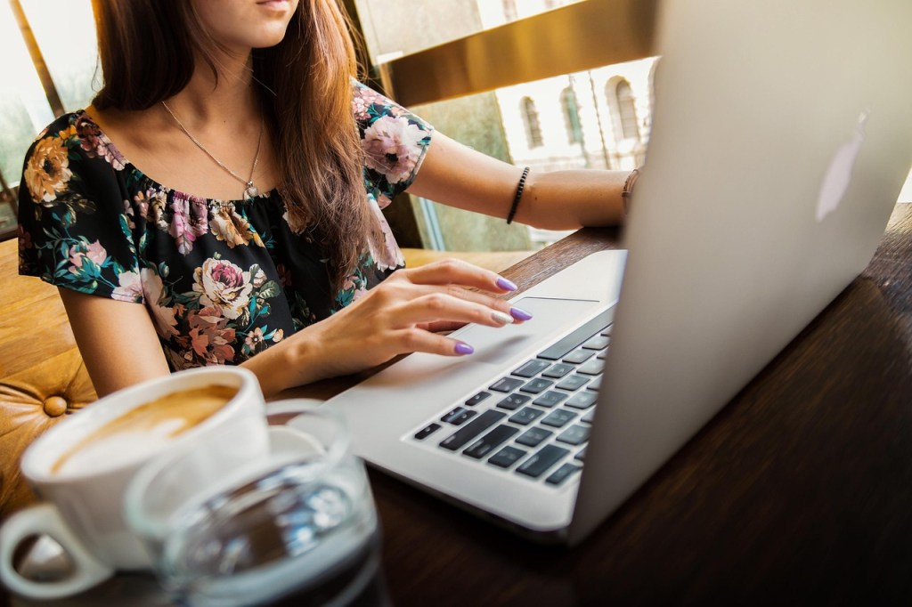 A person typing on a laptop in a cafe, wearing a floral blouse, with a cup of coffee and a glass of water on the table.