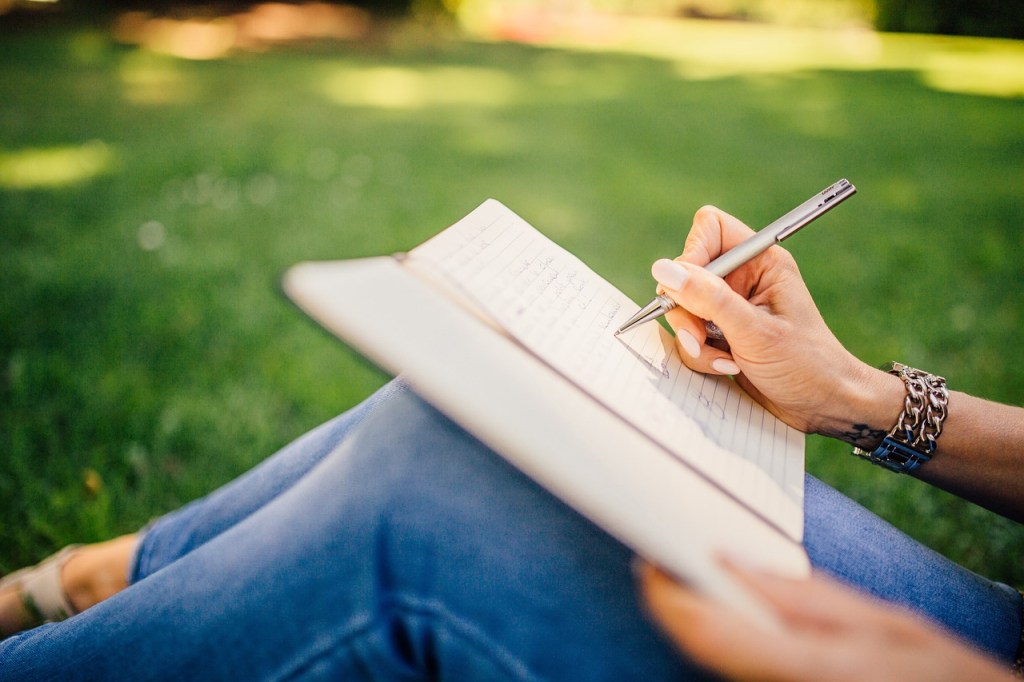 A close-up of a hand holding a pen, writing in a notebook while seated on grass, indicating a moment of reflection and journaling.