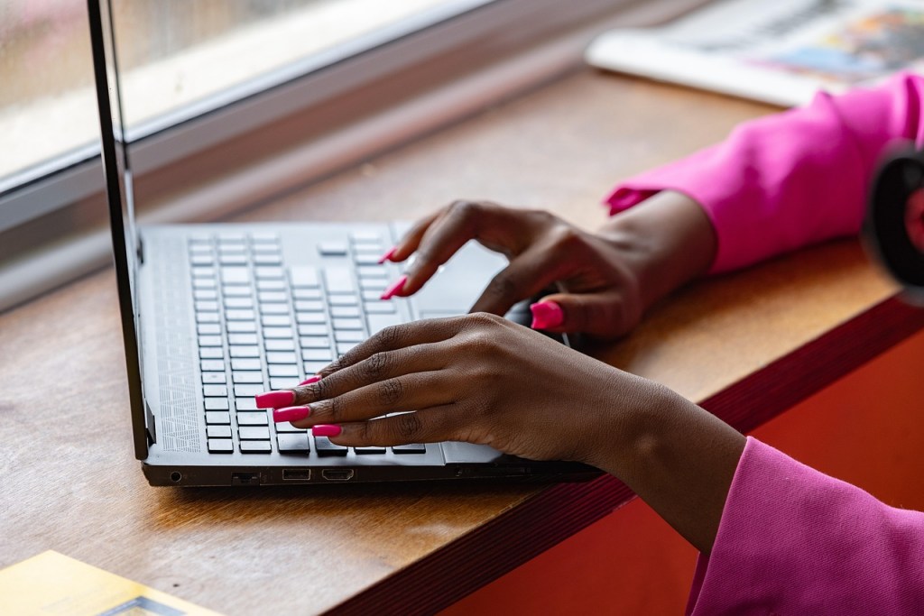 Close-up of hands typing on a laptop keyboard, with vibrant pink nails, resting on a wooden surface near a window.