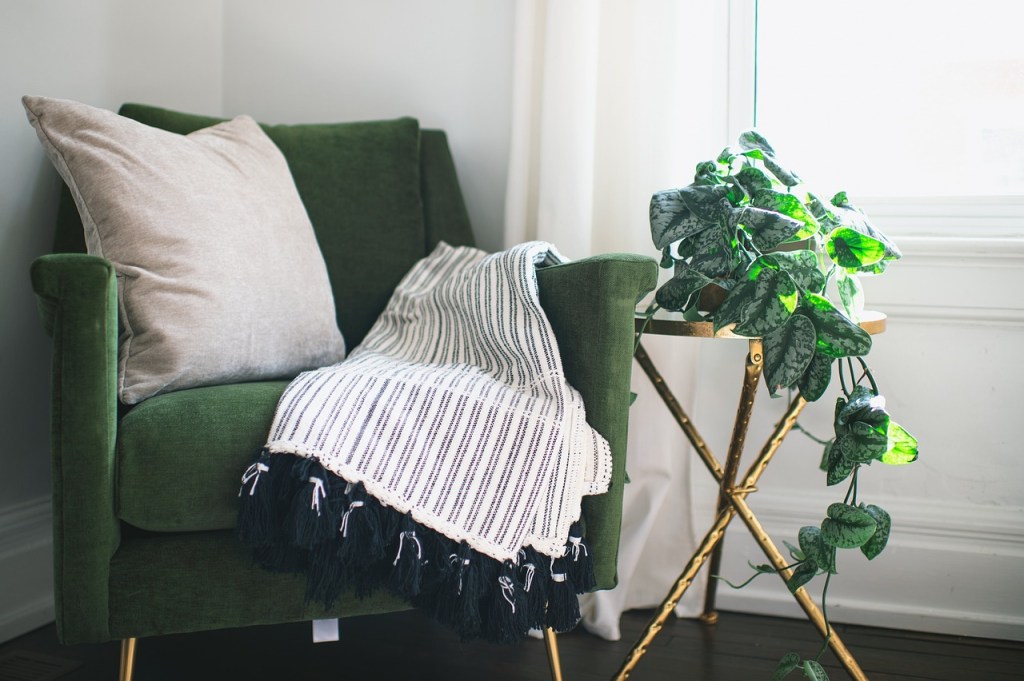 A cozy green armchair with a beige pillow, draped with a striped blanket featuring tassels, next to a gold side table adorned with lush, green foliage.