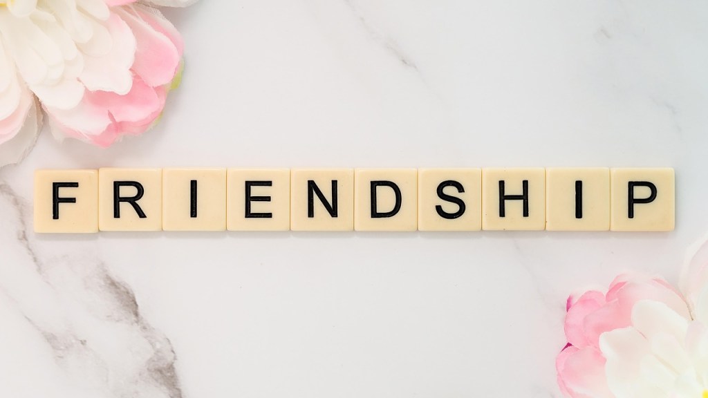 The word 'FRIENDSHIP' spelled out with letter tiles on a marble surface, surrounded by pink flowers.