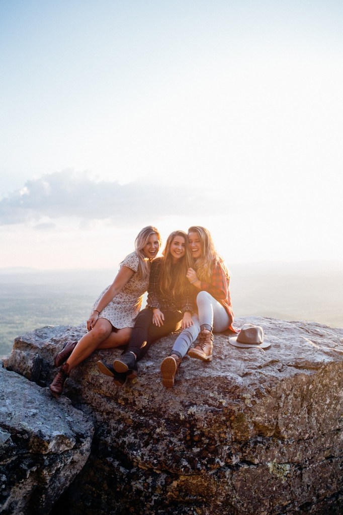 Three young women sitting on a rock ledge, smiling and enjoying each other's company, with a scenic landscape in the background.
