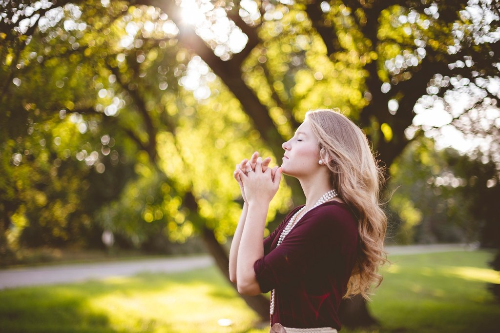 A young woman with long, wavy hair is standing outdoors with her eyes closed, holding her hands together in front of her chest. She appears to be in a reflective or meditative state, surrounded by lush greenery and softly glowing sunlight.