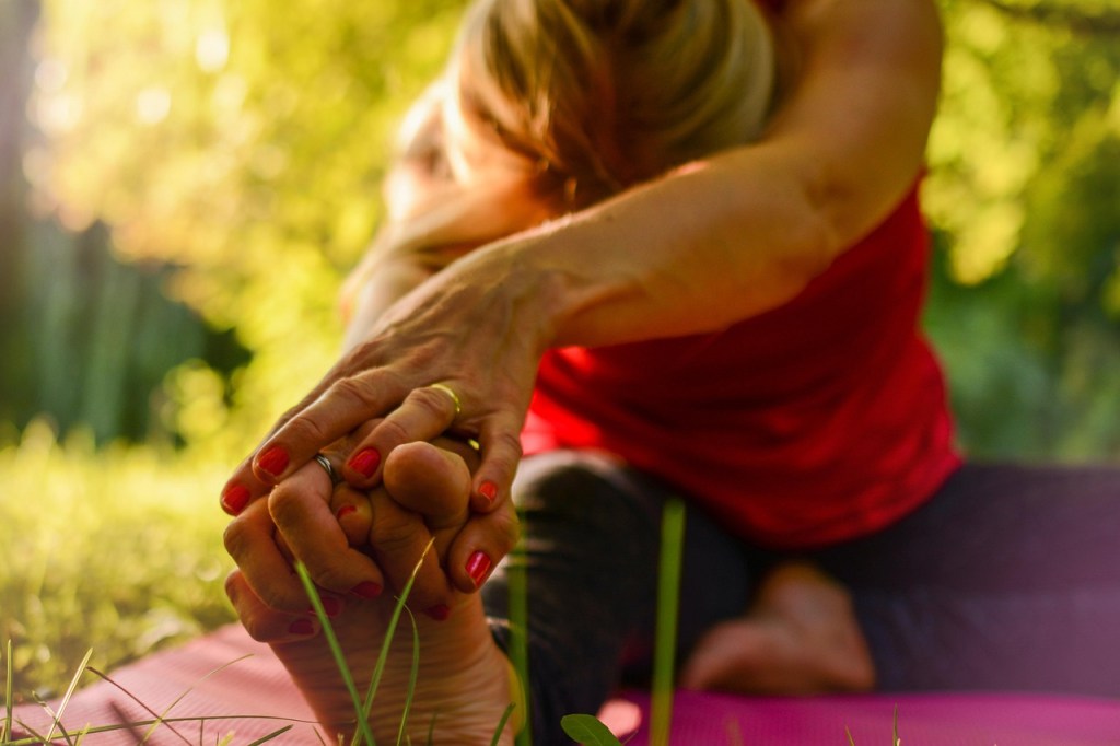 A person practicing yoga outdoors, stretching forward and reaching for their feet, surrounded by greenery and natural light.